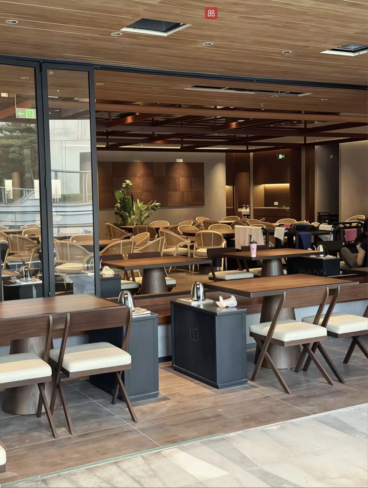 Rectangular restaurant tables with dark-framed cushioned chairs, with lighter curved-back seating visible further inside.